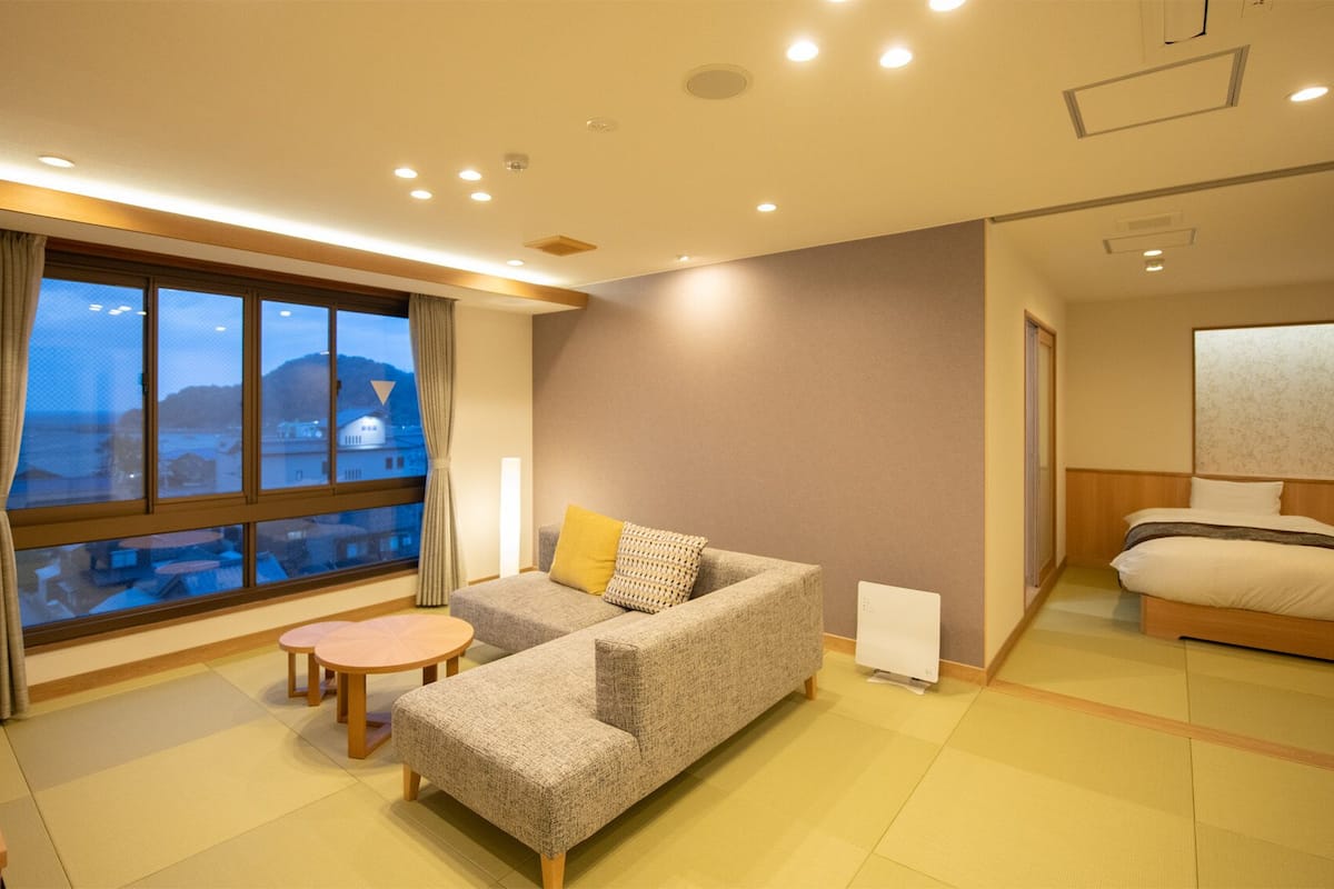 A hotel room with a sofa, a small table, a bed, a large window with a view, and a bedside table with a lamp.