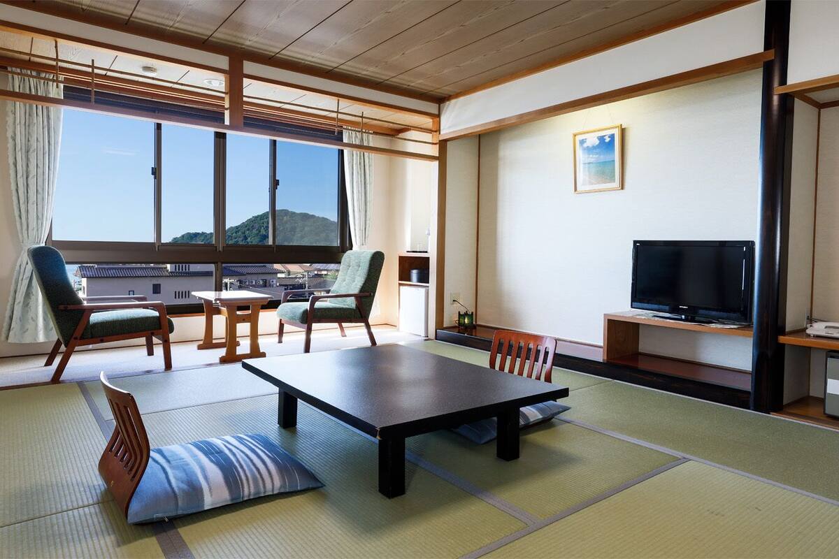 A traditional Japanese room with tatami flooring, a low table, chairs, a television, and a view of a mountain.