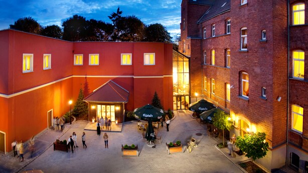 Een binnenplaats met een rood gebouw, een gebouw van baksteen en buitenmeubilair met parasols.