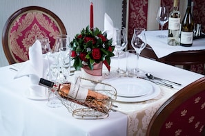 Una mesa de comedor dispuesta con mantel blanco, copas de vino, una vela y un ramillete de rosas rojas.