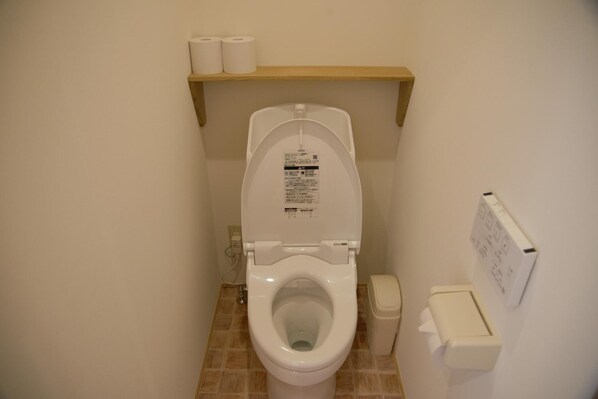 Et standard toilet med hvid skål og låg, toiletpapir på en træhylde over toilettet, og en hvid skraldespand til højre.