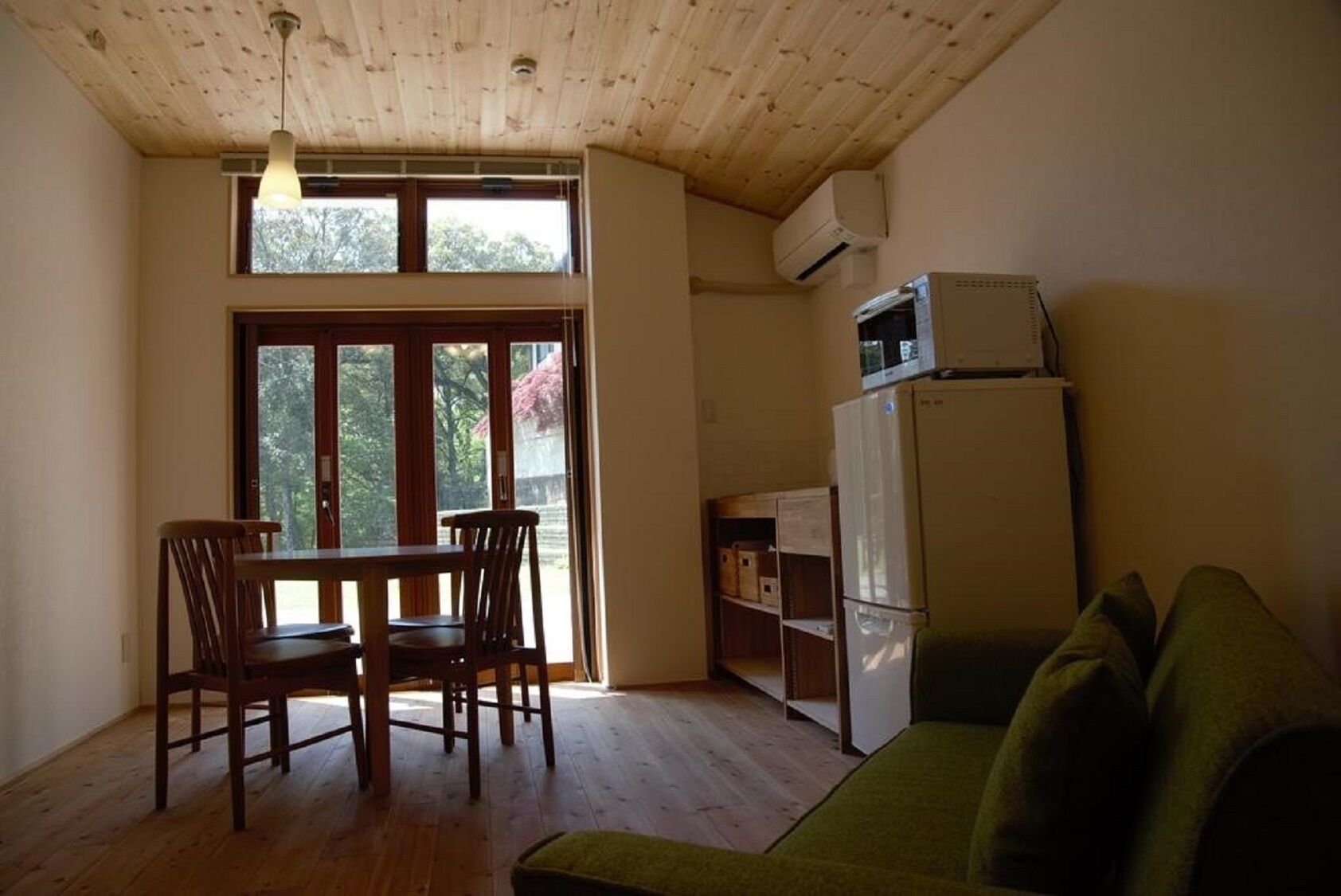 A room with a wooden ceiling, a dining table with chairs, a green sofa, a white refrigerator, and a window with a view of trees.