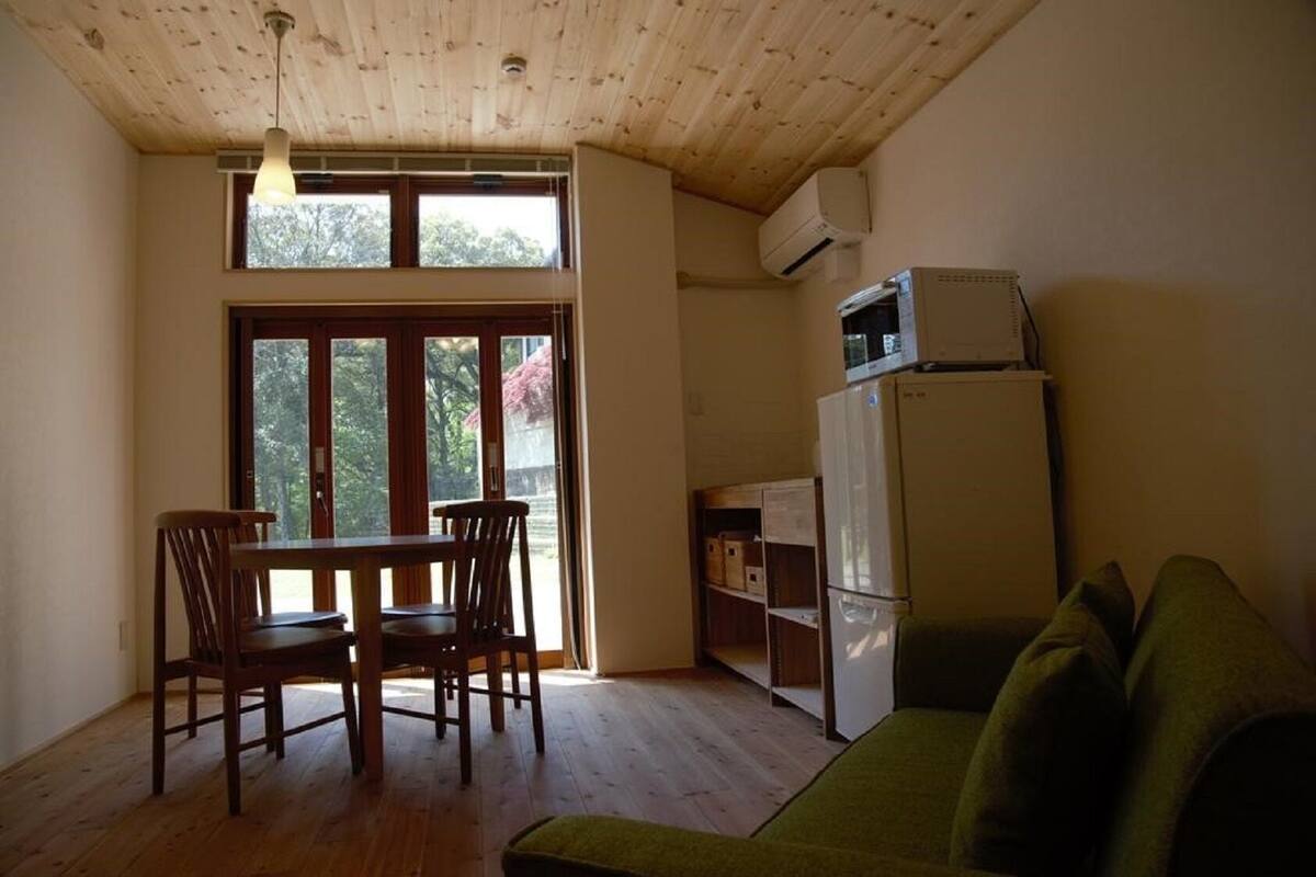 Ein Zimmer mit Holzbalkendecke, einem Esstisch mit Stühlen, einem grünen Sofa, einem weißen Kühlschrank und einem Fenster mit Blick auf Bäume.