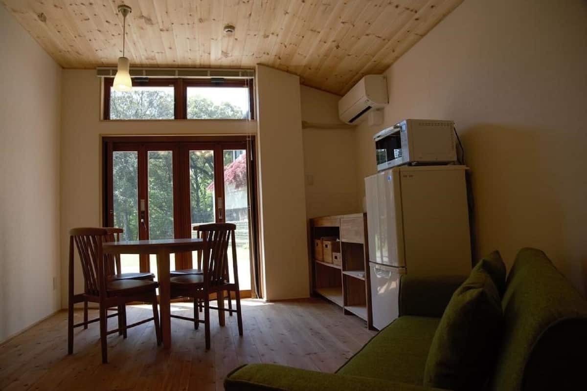 A room with a wooden ceiling, a dining table with chairs, a green sofa, a white refrigerator, and a window with a view of trees.