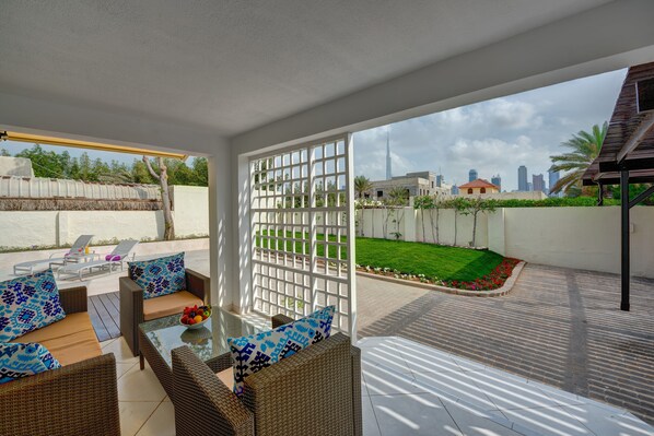 A covered outdoor seating area with wicker furniture and a glass table, overlooking a well-maintained garden and city skyline.