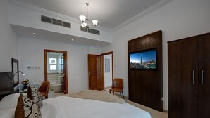 A bedroom with a large bed, a TV mounted on a wooden cabinet, a chair, and a bathroom visible through an open door. - J5 Four Bedroom Villa in Mirdif (Dubai)