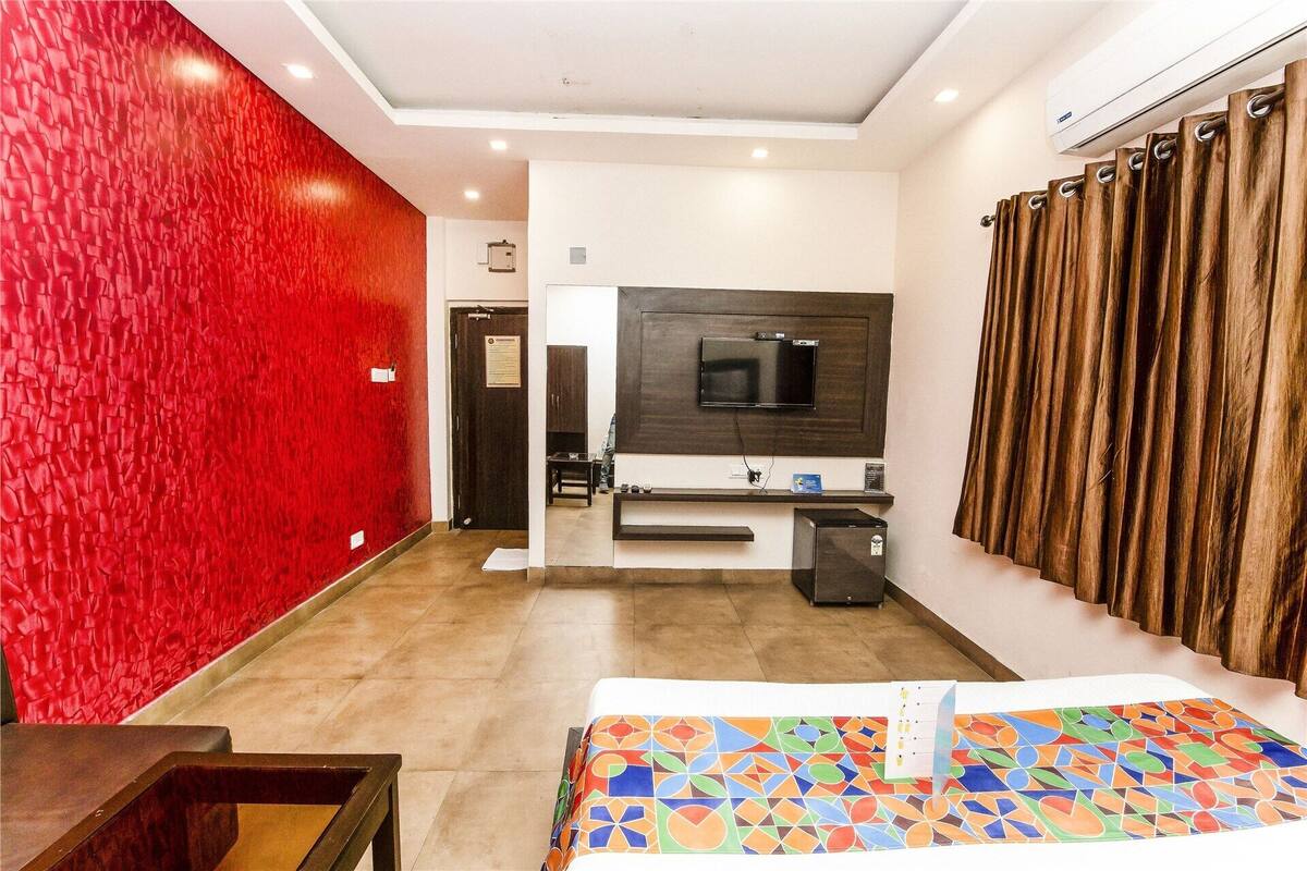Ein Hotelzimmer mit roher, roter Wand, einem an einer dunklen Holzvertäfelung montierten Fernseher, einem Bett mit farbenfroher, geometrischer Tagesdecke und einem Fenster mit braunen Vorhängen.