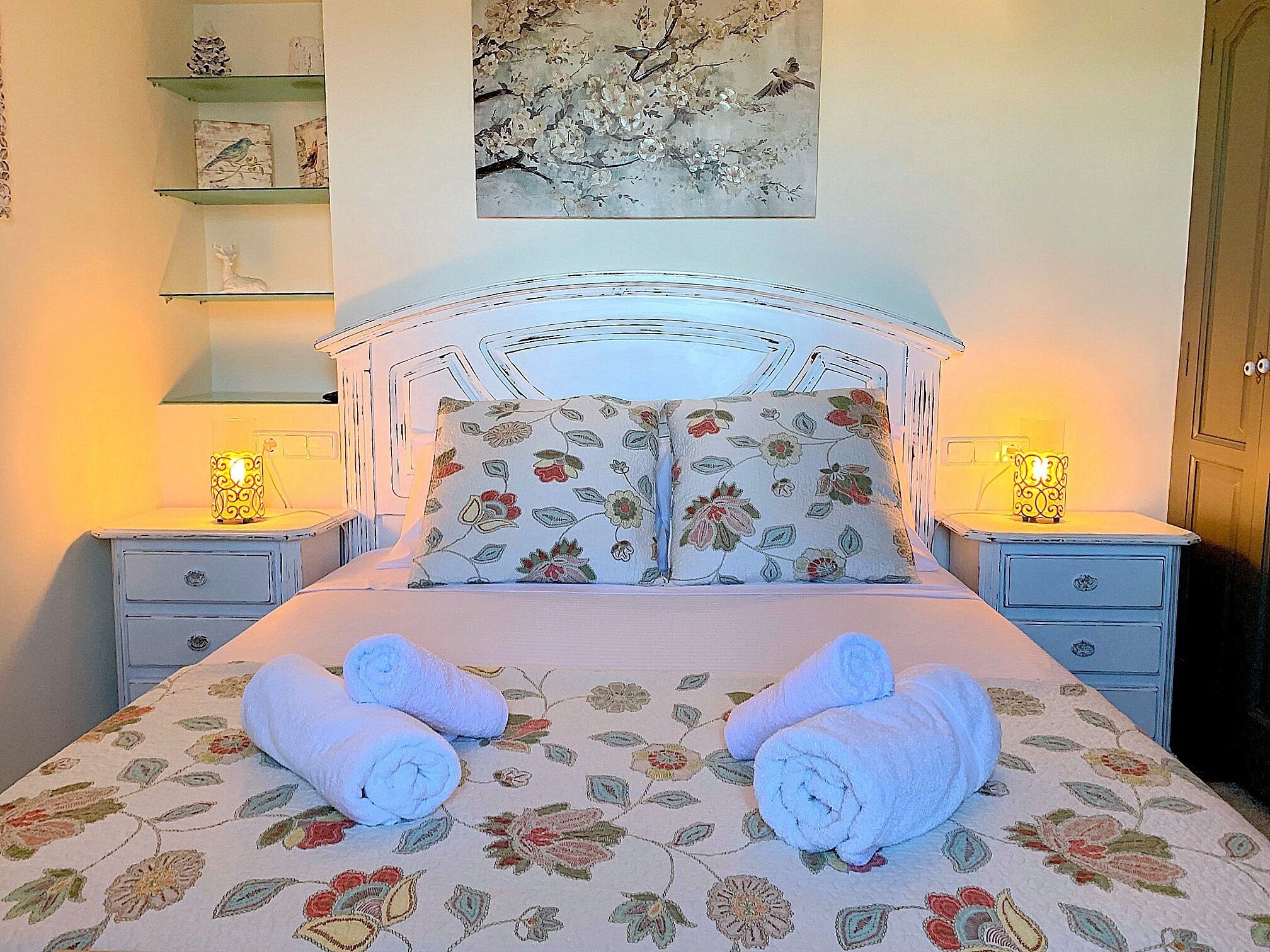 A bed with floral bedding, two pillows, and two rolled towels. There are bedside tables with lamps and a wall-mounted shelf with decorative items.