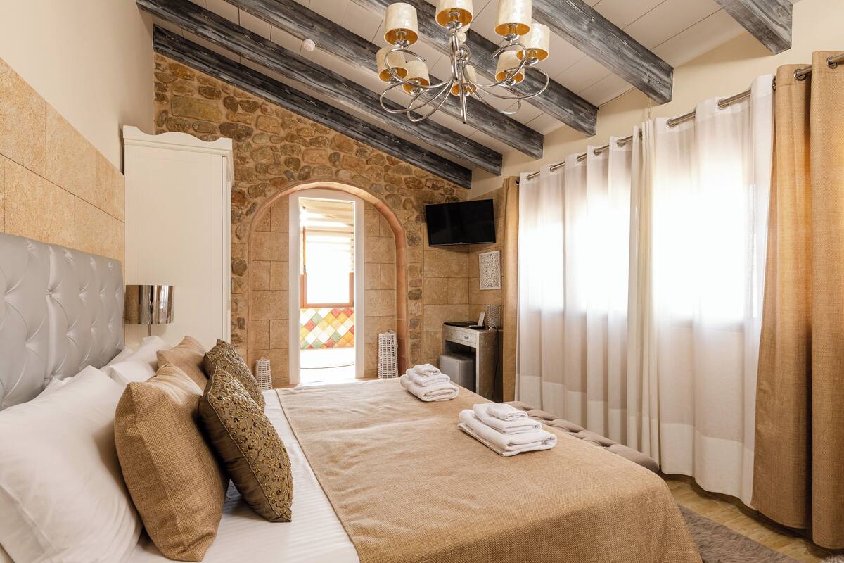 A bedroom with a stone wall, a bed with beige bedding, a chandelier, and a TV mounted on the wall.