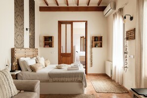 Un dormitorio con cama, mesitas de noche, cabecera de madera, lámpara de noche, estante en la pared y ventana con cortinas.