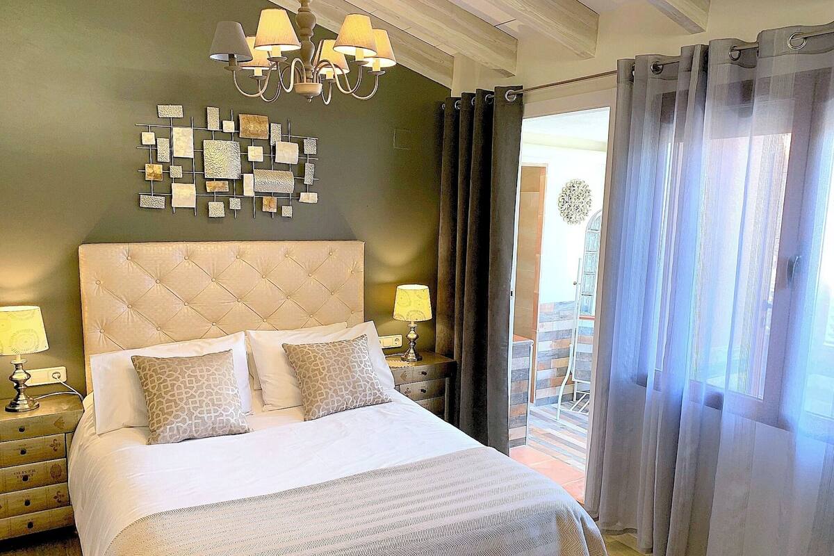 A bedroom with a large bed, two bedside tables with lamps, a decorative wall piece, and a view through sheer curtains.