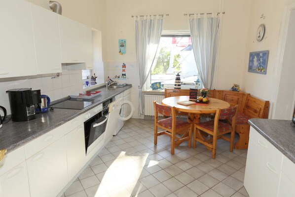 Une cuisine avec des meubles blancs, un four encastré, un lave-linge, une table à manger avec des chaises et une fenêtre avec des rideaux.