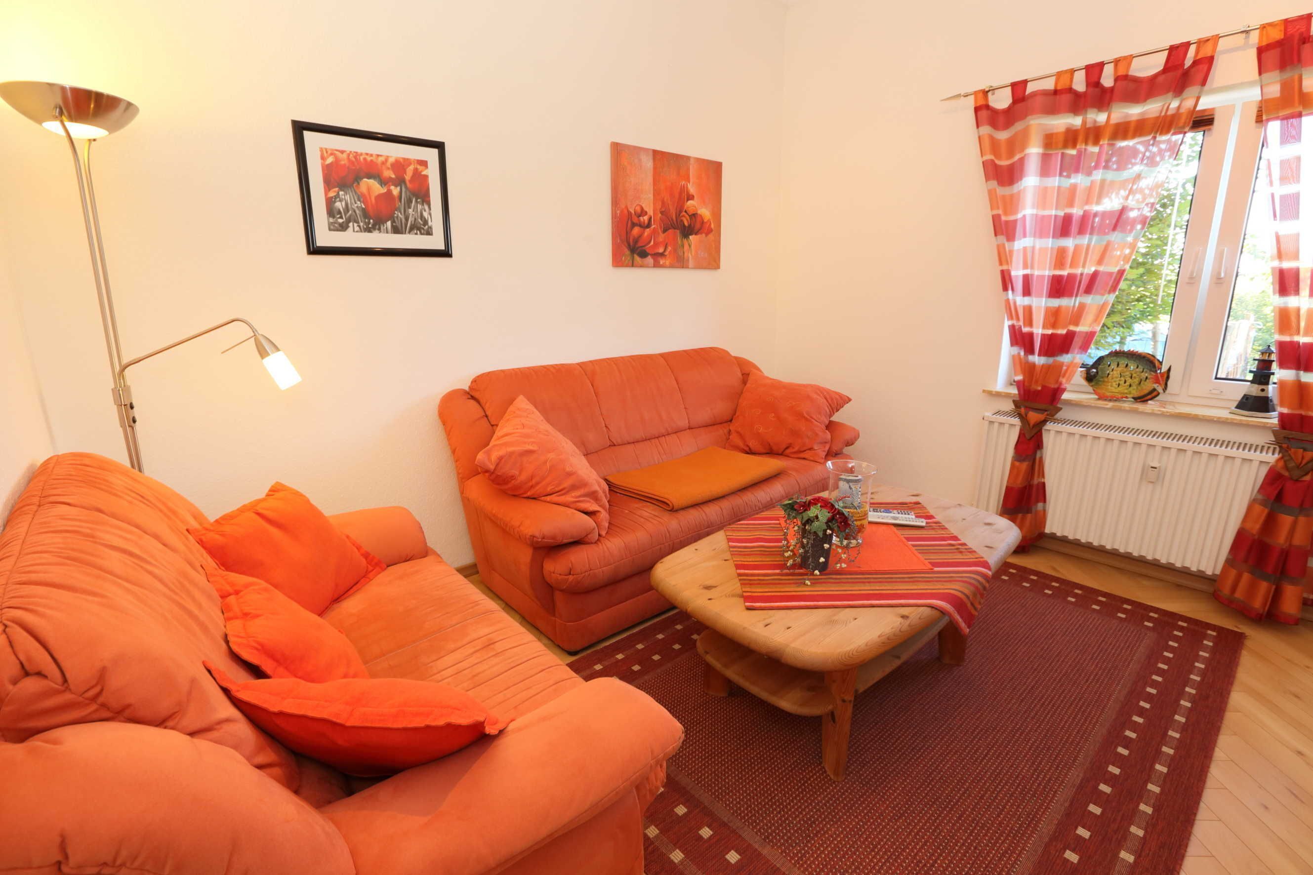 A living room with an orange sofa, a wooden coffee table, and a floor lamp. There are two framed pictures on the wall.