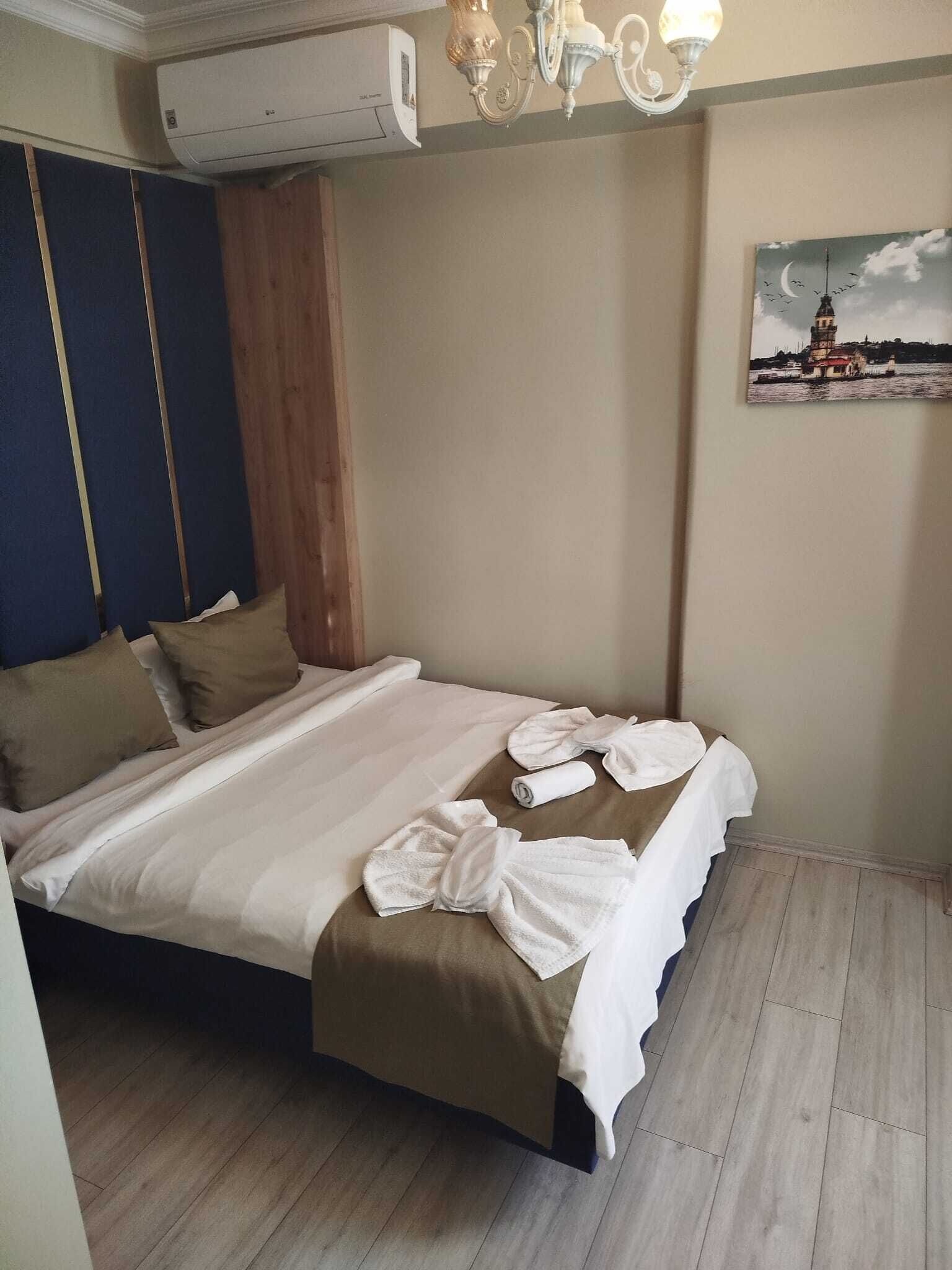 A hotel room with a bed, pillows, a towel, an air conditioning unit, a chandelier, and a painting on the wall.
