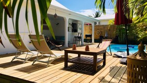 En træterrasse med liggestole og et bord, en pool og et overdækket område med en bar.