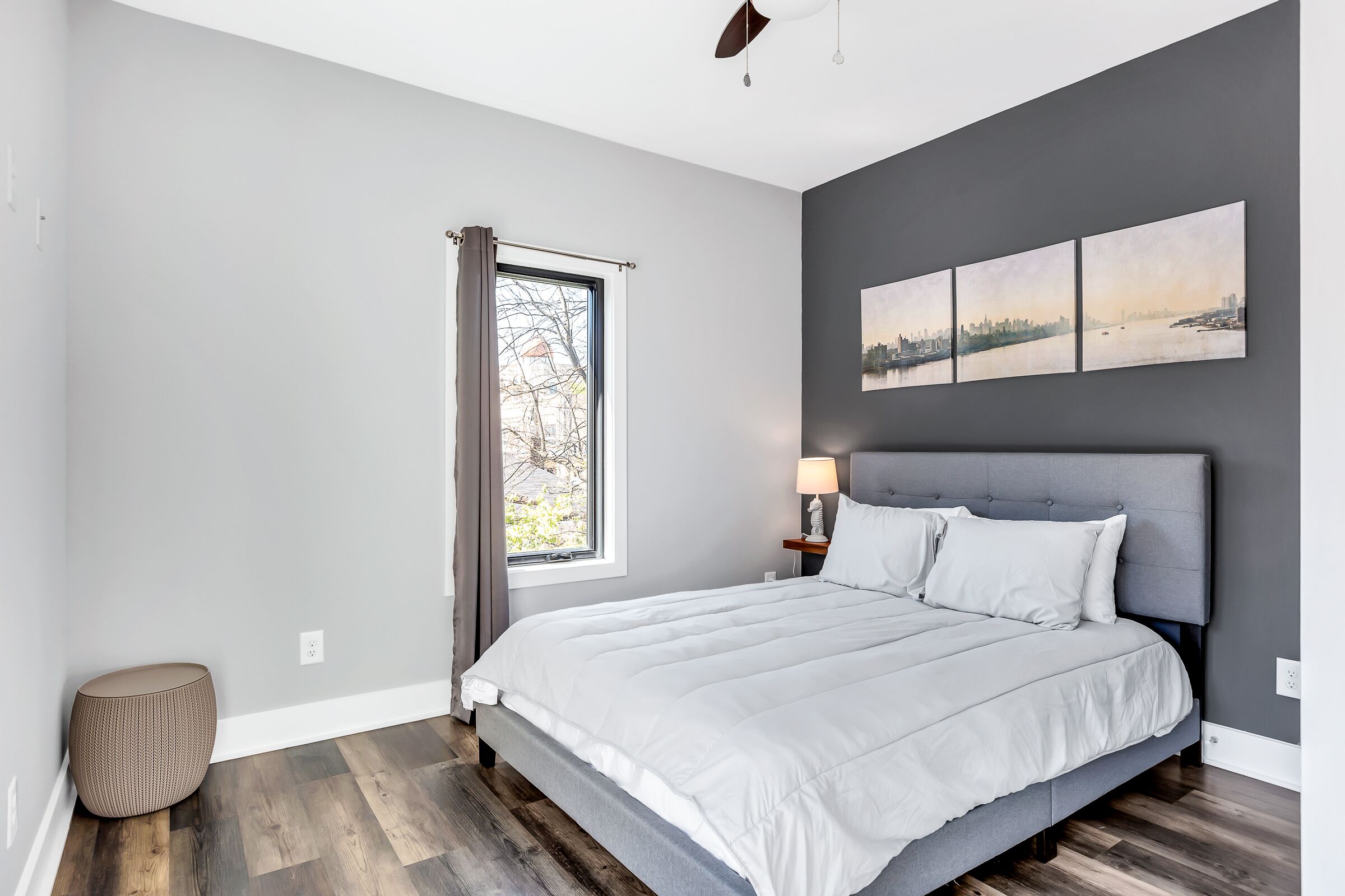 A bedroom with a bed, a window with curtains, a nightstand with a lamp, and a cityscape painting on the wall.