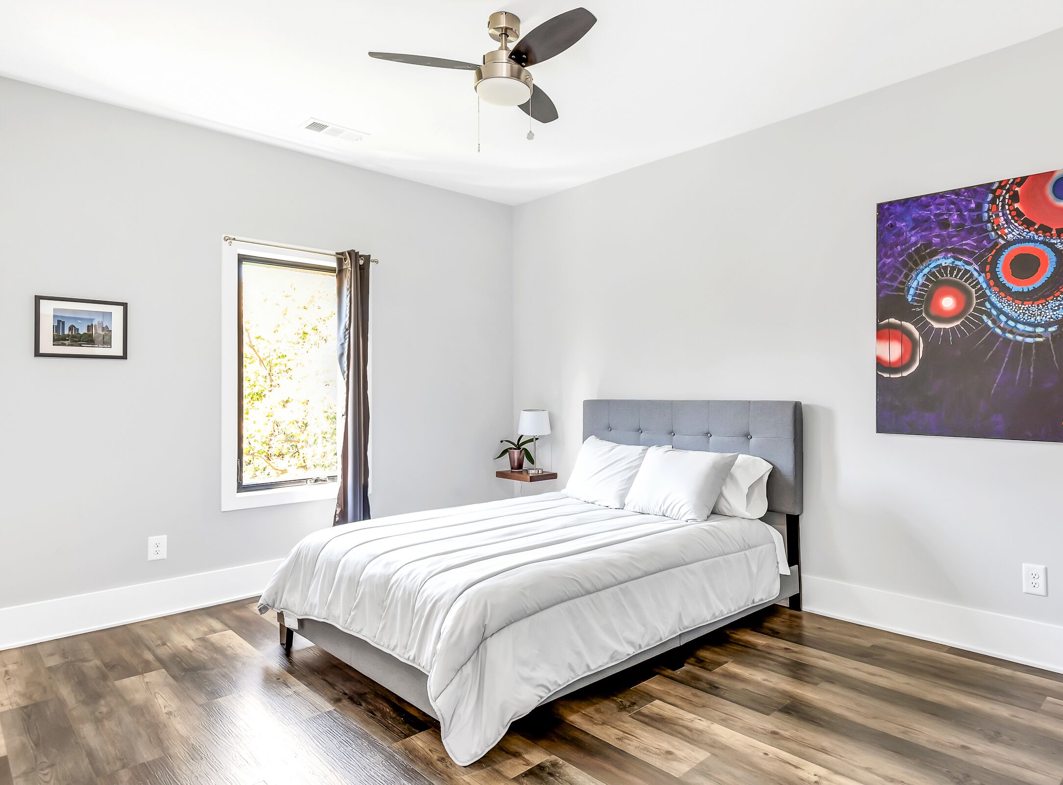Une chambre à coucher avec un lit, un ventilateur de plafond, une fenêtre avec des rideaux, une table de chevet avec une lampe et un mur orné d’un tableau.