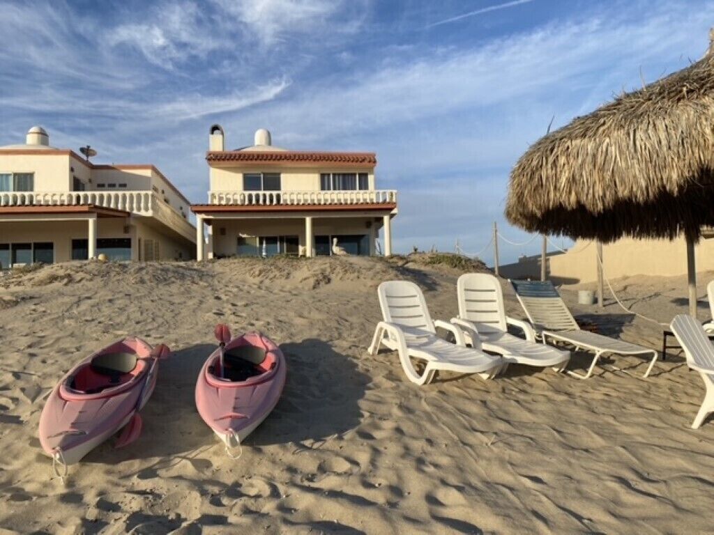 Une plage avec des chaises longues et un parasol en chaume, un bâtiment doté de balcons, et deux bateaux.