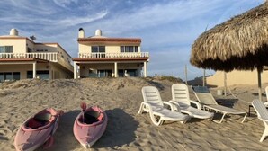 Une plage avec des chaises longues et un parasol en chaume, un bâtiment doté de balcons, et deux bateaux.