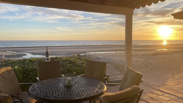 Un espace extérieur abrité, avec une table et des chaises, donnant sur une plage où l’on peut admirer le coucher du soleil.