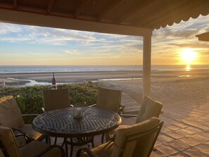 Un espace extérieur abrité, avec une table et des chaises, donnant sur une plage où l’on peut admirer le coucher du soleil.