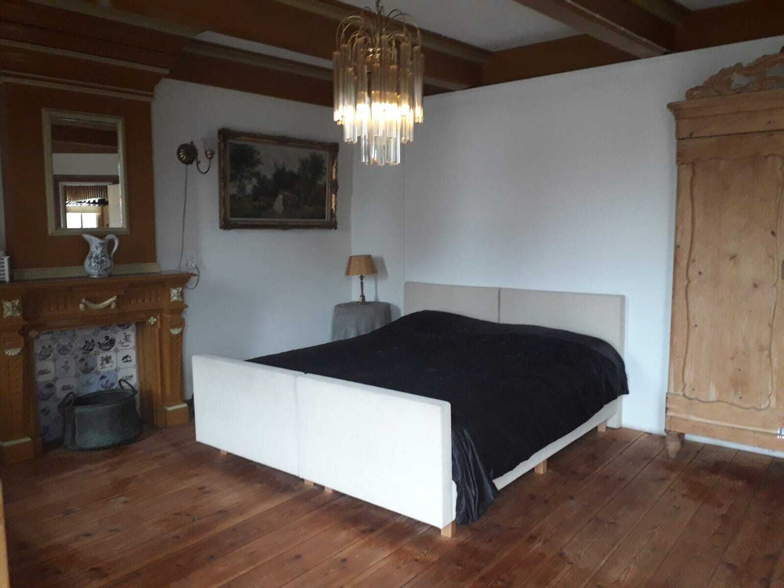 Une chambre avec un plancher en bois, un lit blanc, une armoire en bois, une cheminée et un lustre.