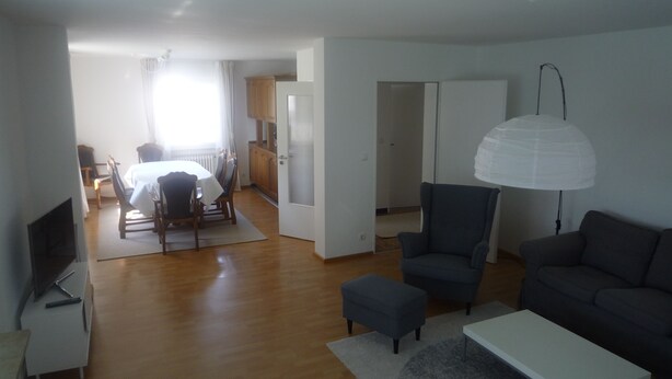 A modern living room with a grey sofa, a white coffee table, and a blue armchair. There is a dining area with a table and chairs in the background.