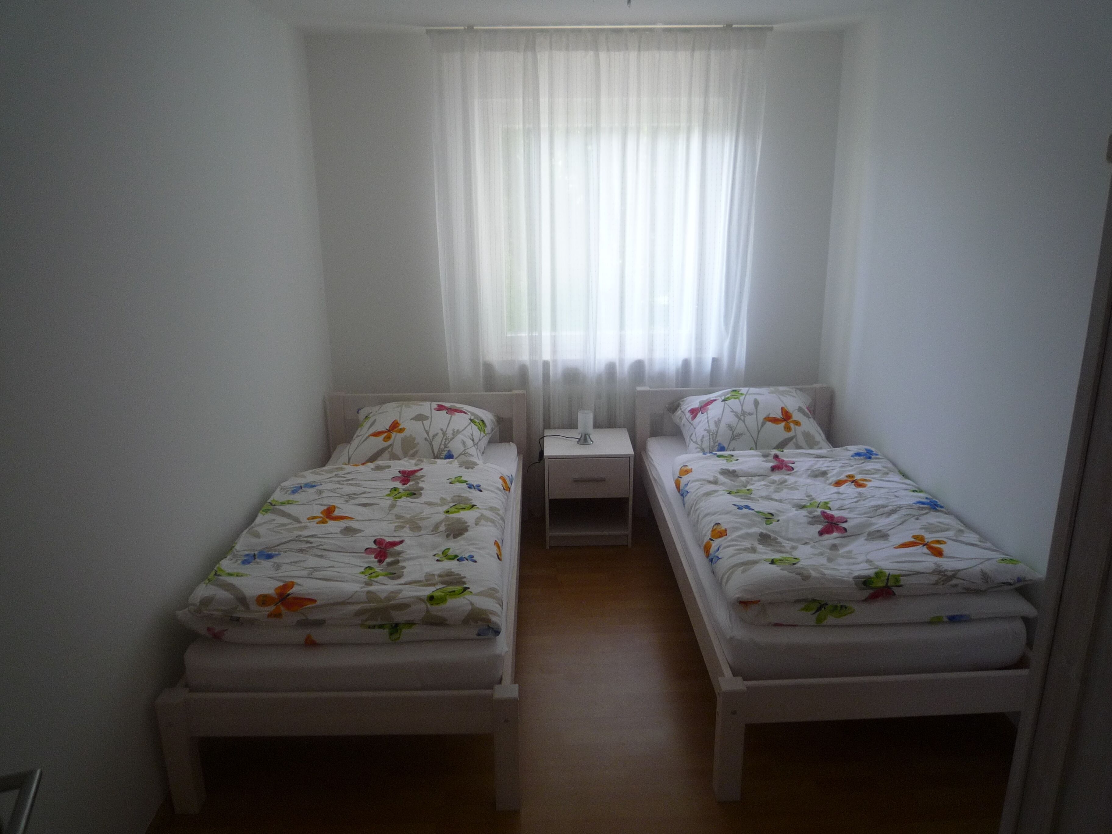Une petite chambre propre, avec deux lits simples, une table de chevet et une fenêtre ornée de rideaux légers.
