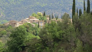 Un village perché sur une colline, avec des bâtiments traditionnels en pierre, une végétation luxuriante et de grands cyprès.