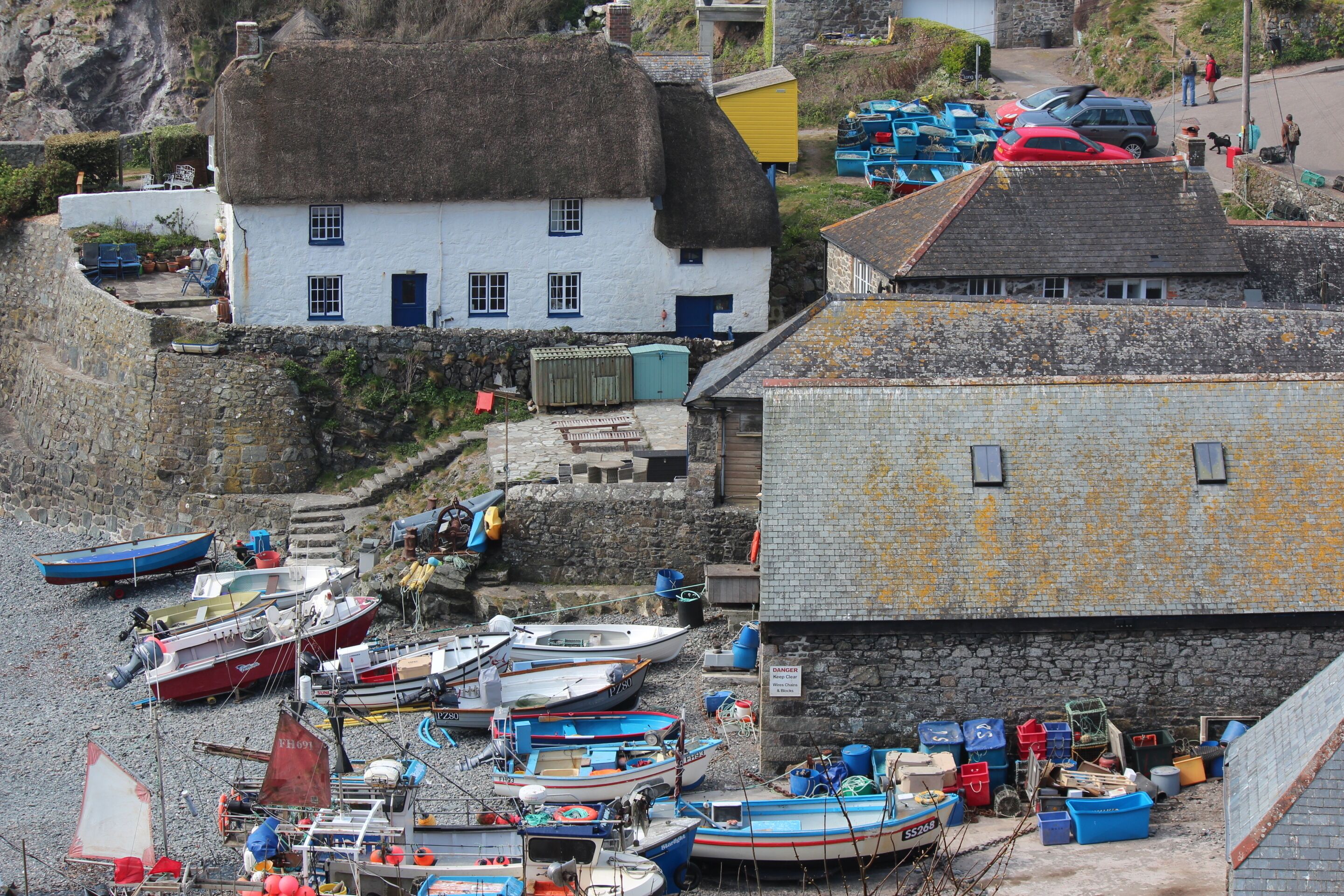 A coastal village with thatched-roof houses, a stone wall, and a variety of boats docked along the shore.