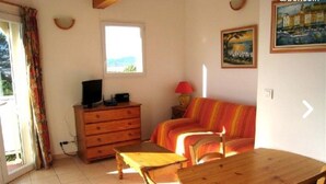 A room with a wooden table and chairs, a sofa, a TV on a wooden cabinet, a window with a view, and a lamp. - Studio 25 M2, air-conditioned 350mt from the beach. (Hyères)
