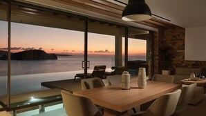 A modern dining area with a wooden table and chairs, large windows offering a view of the sea and sunset, and a hanging pendant light. - Alkyonides Villas (Zakynthos)