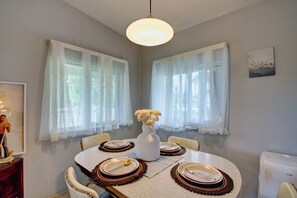 Een eetkamer met een ronde tafel voor vier personen, witte gordijnen, een witte vaas met bloemen en een schilderij aan de muur.