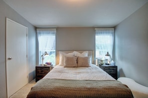 Un dormitorio con una cama, dos mesitas de noche, una mesita auxiliar, una puerta y una ventana con cortinas.