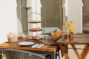 Una mesa de desayuno con pan, bollería y una bandeja de fruta, además de un botellón de zumo de naranja y vasos.