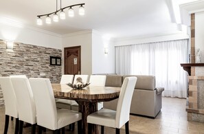 Un comedor con mesa de madera y sillas blancas, pared de piedra, puerta de madera y un sofá al fondo.