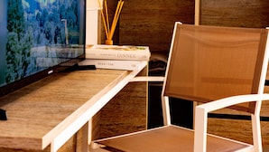 Una habitación con televisión mostrando un paisaje urbano, un escritorio de madera con una silla, y un armario con ropa colgada.