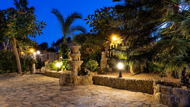 Un patio empedrado con plantas tropicales y estructuras de piedra, iluminado con luz ambiental.