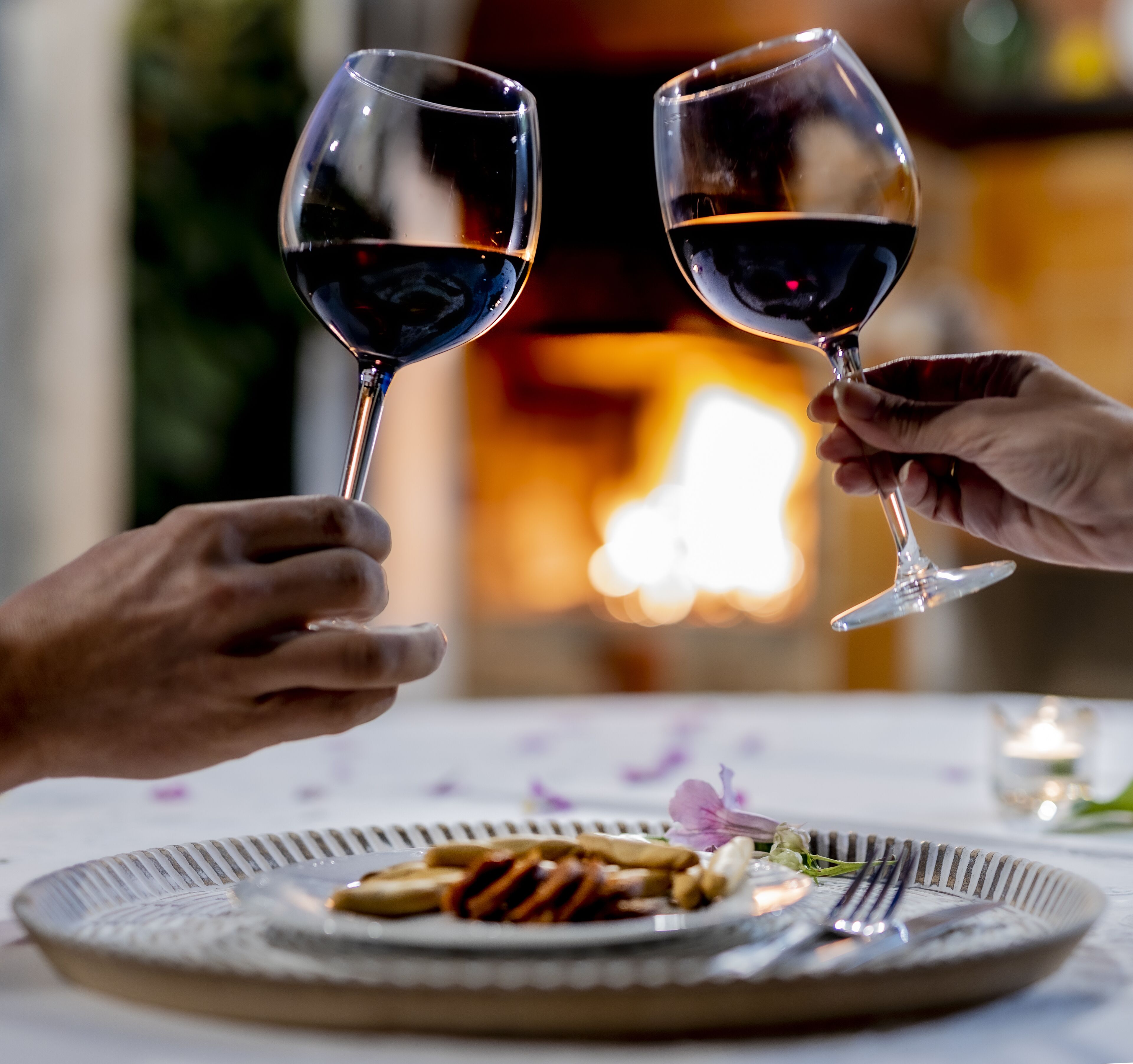 Two people toasting with wine glasses at a table with a plate of food and a candle.