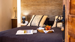 Una cama bien hecha con un edredón oscuro, dos almohadas estampadas, una mesita de noche con una lámpara, un pequeño jarrón con flores y una cesta de fruta sobre la cama.
