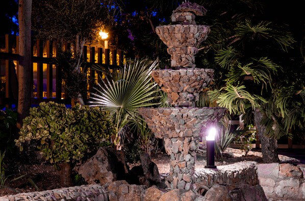 Un jardín nocturno con una fuente de piedra, plantas de palma y una cerca de madera al fondo.