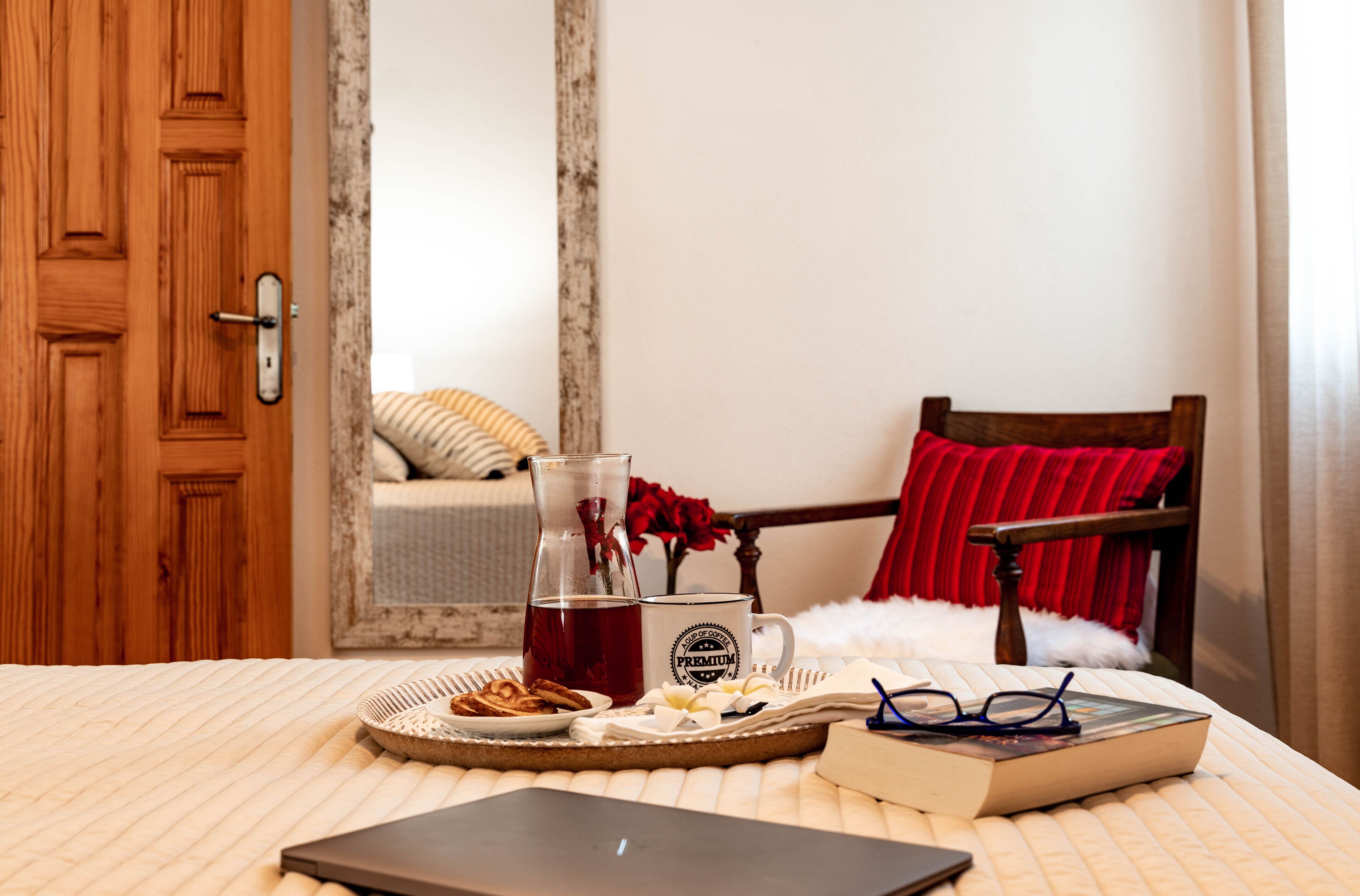 A neatly arranged breakfast tray with a beverage, a book, glasses, and a laptop on a bed with a striped bedspread.