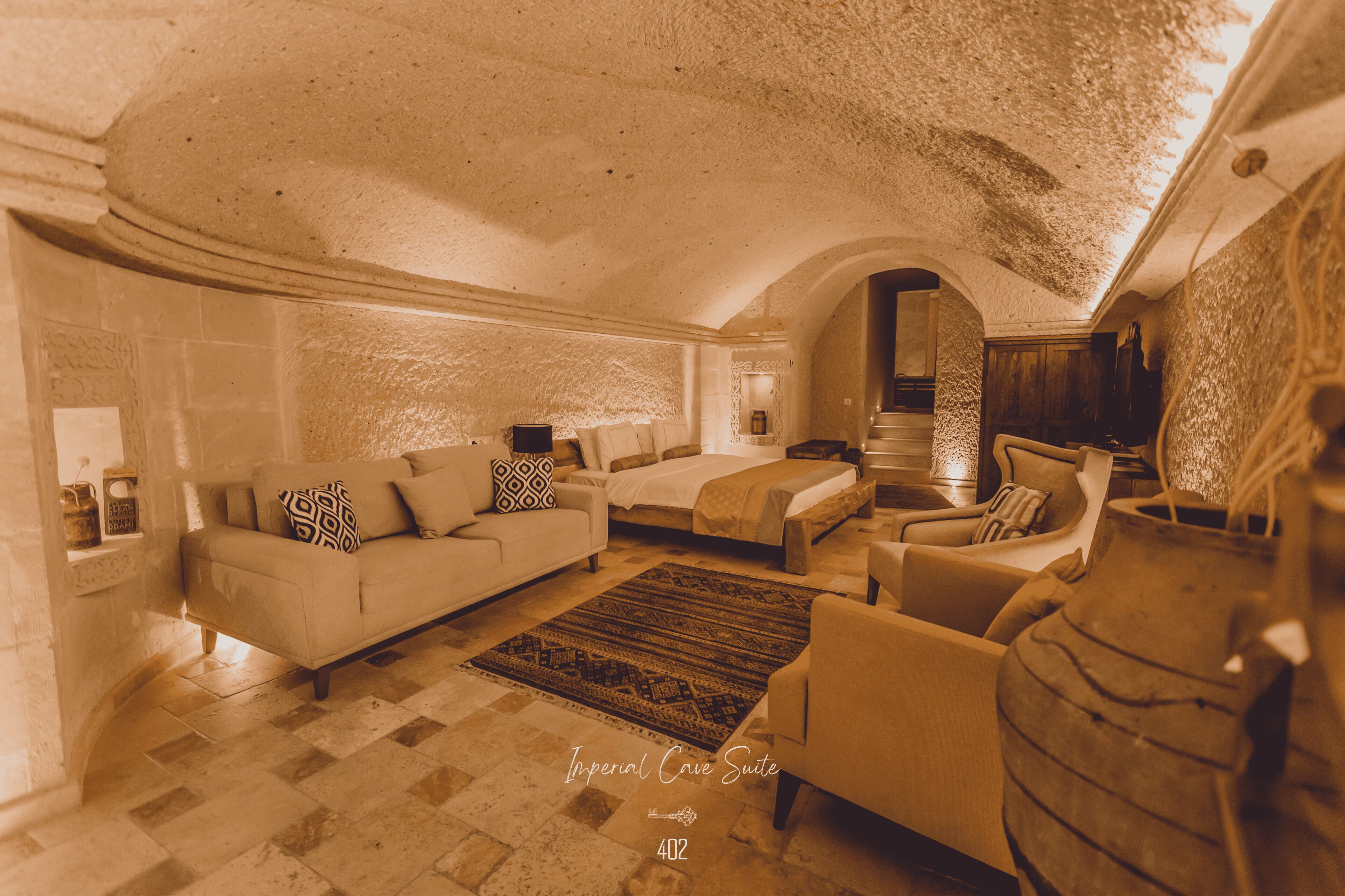 A spacious suite with a large bed, a sofa, and a chair, featuring a textured ceiling and stone walls.