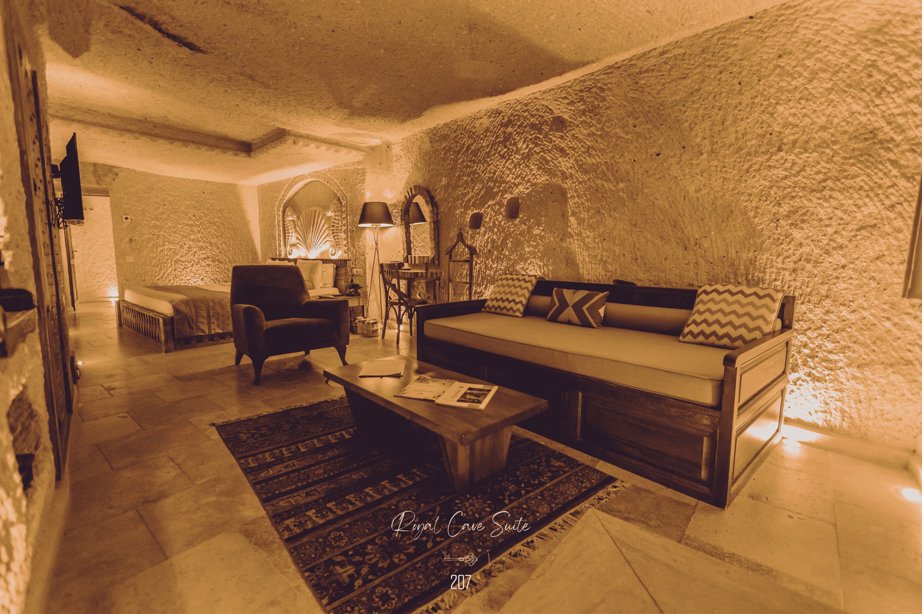 A cozy living room with a sofa, armchair, and coffee table, featuring a rustic stone wall and a patterned rug.