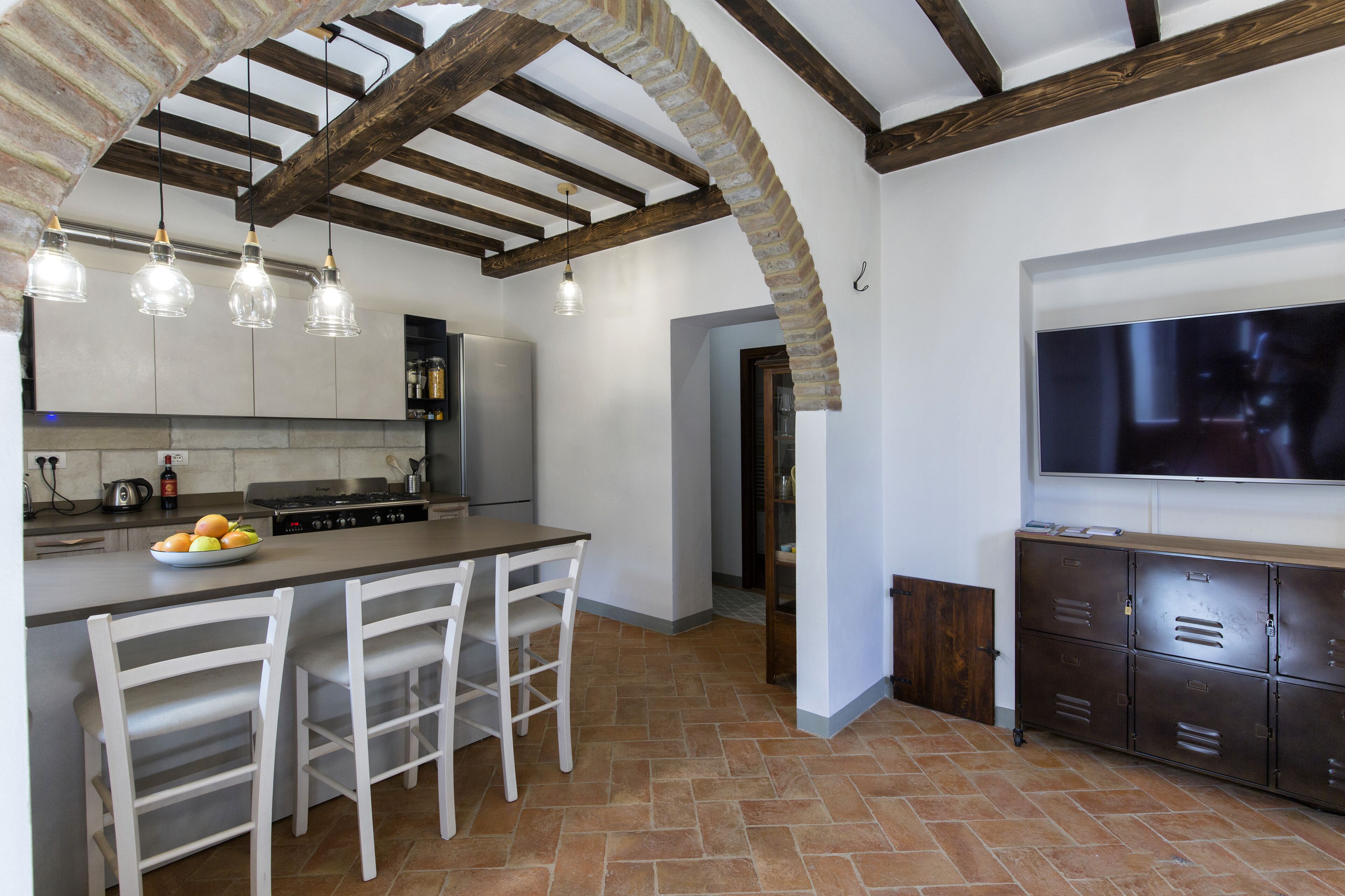 Una cocina con una isla central, sillas blancas, un televisor montado en la pared y un arco de piedra.