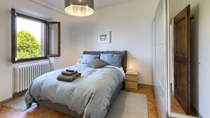 Ein Schlafzimmer mit Bett, Nachttisch, Fenster, Heizkörper und einer Tür.
