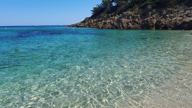 Uma praia clara com águas rasas, falésias rochosas e vegetação escassa.