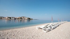 A pebbled beach with rows of white lounge chairs and striped umbrellas, a clear blue sky, and buildings in the background. - Luxurious Penthouse with Wonderful Sea and Old Town View (Primosten)