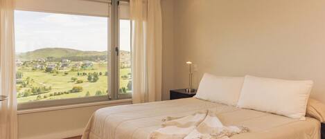 A bedroom with a large bed, a nightstand, a lamp, and a window with a view of a green landscape.