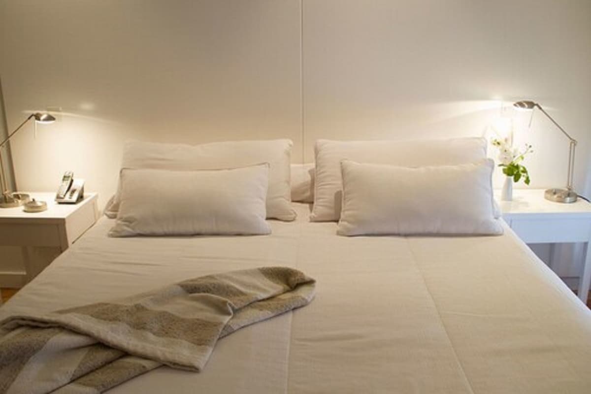 A double bed with white linens and pillows, a bedside table with a lamp, and a blanket on the bed.